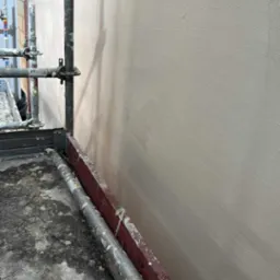 Świeżo nałożony tynk na ścianie, widoczny z bliska ze stojącego rusztowania, z delikatną fakturą i subtelnym oświetleniem.