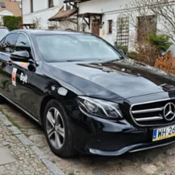 Czarny Mercedes z logo Bolt na drzwiach, zaparkowany na brukowanej ulicy przed białym budynkiem z brązowym dachem i zielenią w tle. Widoczna tablica rejestracyjna.