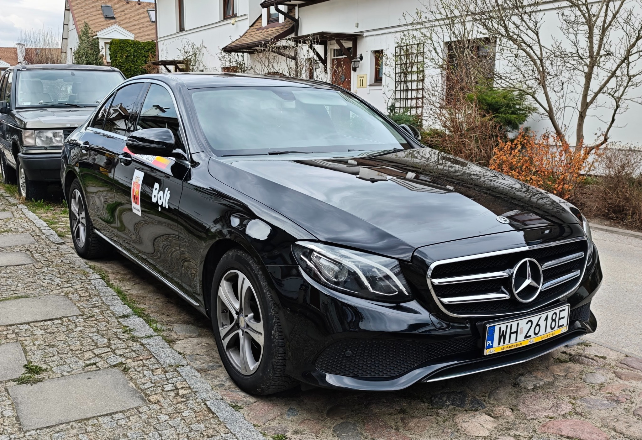 Czarny Mercedes z logo Bolt na drzwiach, zaparkowany na brukowanej ulicy przed białym budynkiem z brązowym dachem i zielenią w tle. Widoczna tablica rejestracyjna.