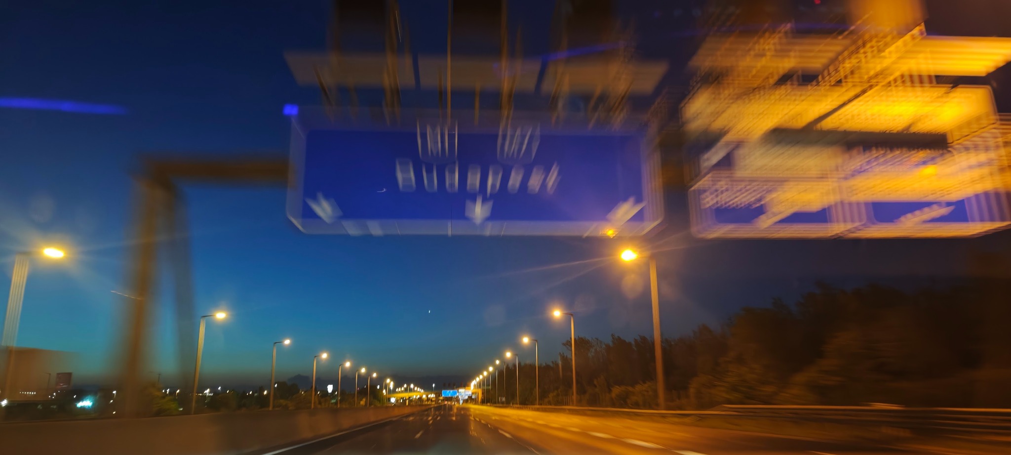 Nocna autostrada z tablicą drogową wskazującą kierunek na Budapeszt, oświetlona przez rzędy lamp ulicznych. Rozmycie ruchu dodaje dynamiki ujęciu.