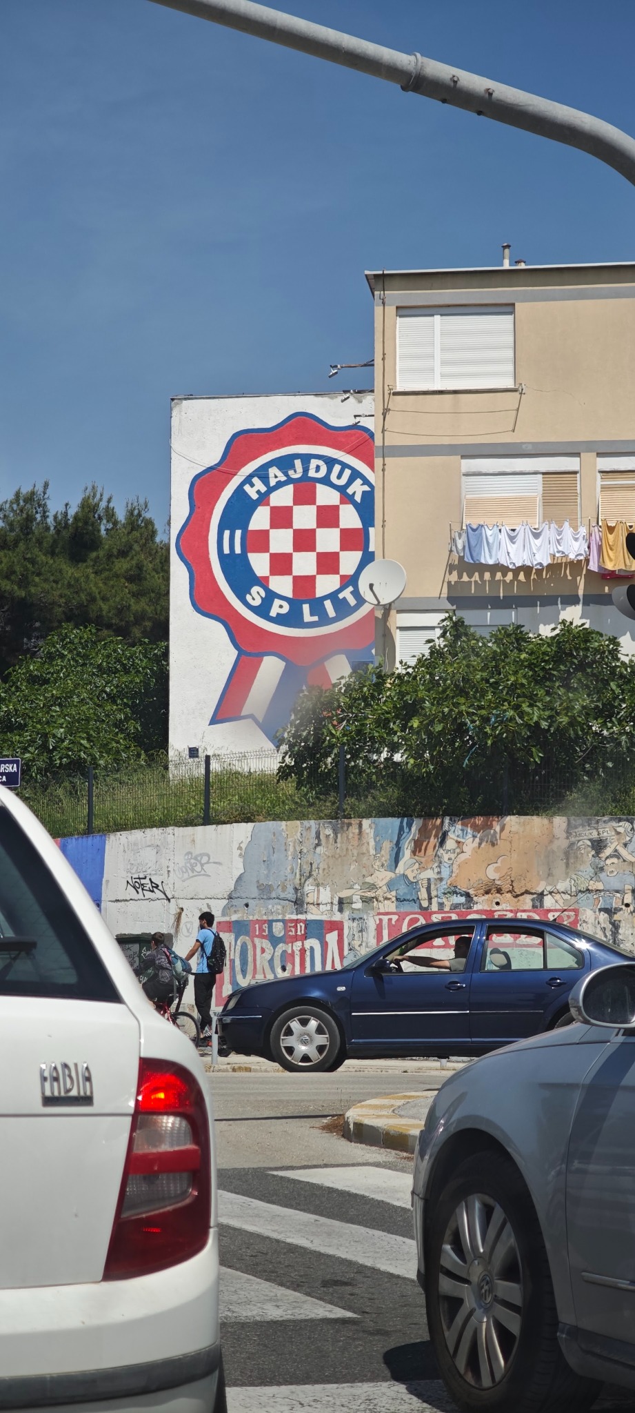 Mural Hajduk Split na ścianie budynku, widoczny z ulicy z przejeżdżającymi samochodami, pieszy na przejściu. Słoneczny dzień, miejski krajobraz.