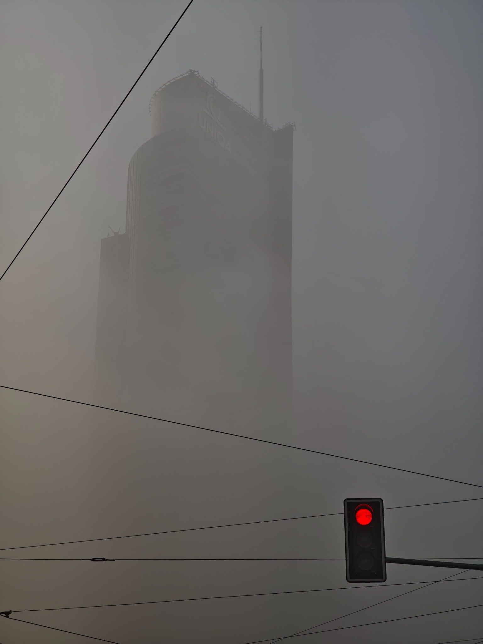 Wieżowiec UNIQA w gęstej mgle, widziany z perspektywy ulicy z czerwonym światłem sygnalizacji świetlnej. Przewody elektryczne przecinają kadr.