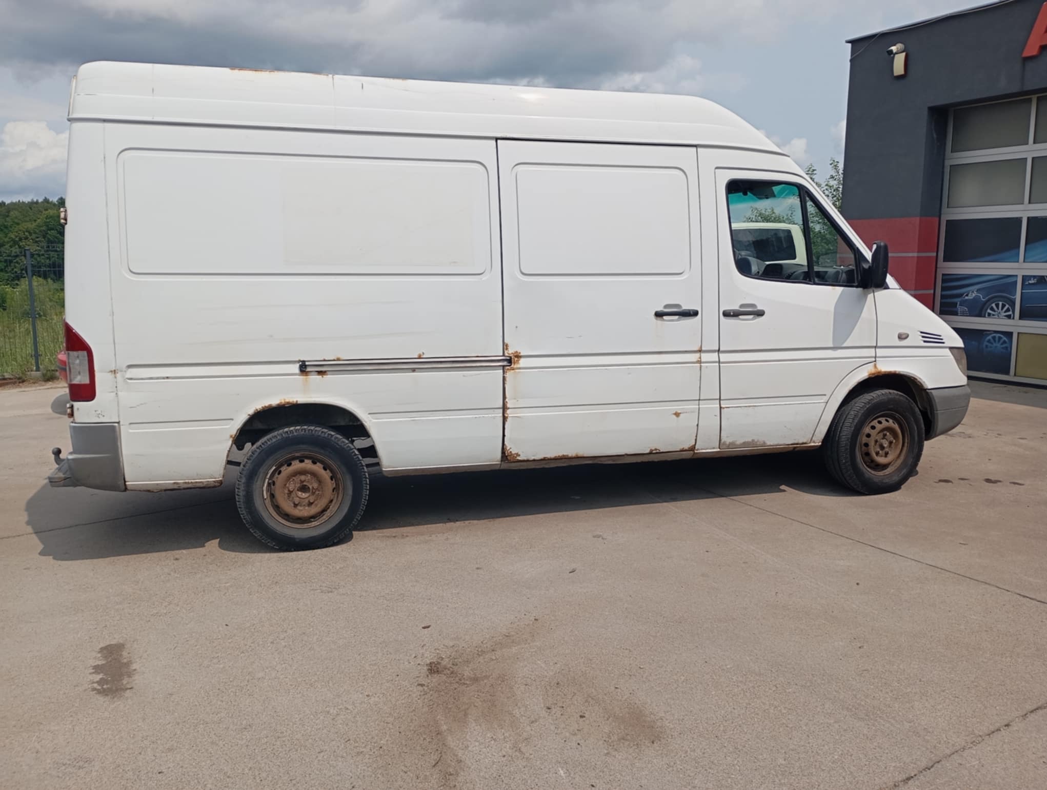Biały, lekko zardzewiały bus dostawczy zaparkowany na betonowym placu, widziany z boku, z budynkiem w tle.