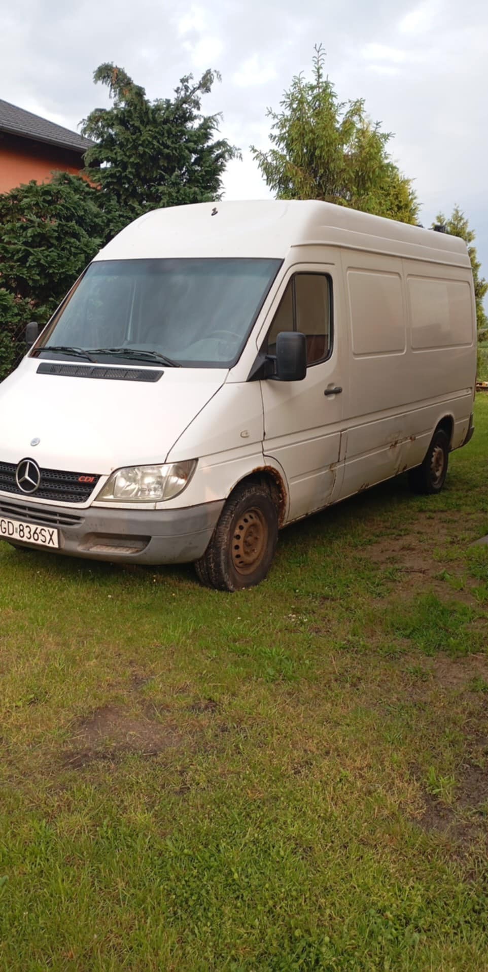 Biały bus Mercedes-Benz Sprinter z widocznymi śladami użytkowania, zaparkowany na trawniku przed budynkiem. Widoczna tablica rejestracyjna i logo marki.