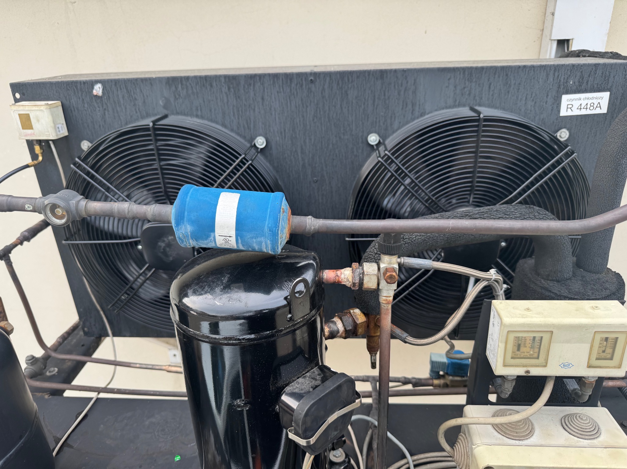 Agregat chłodniczy z dwoma wentylatorami i filtrem, widoczne rury miedziane i manometry. Urządzenie klimatyzacyjne z naklejką 'czynnik chłodniczy R 448A'.