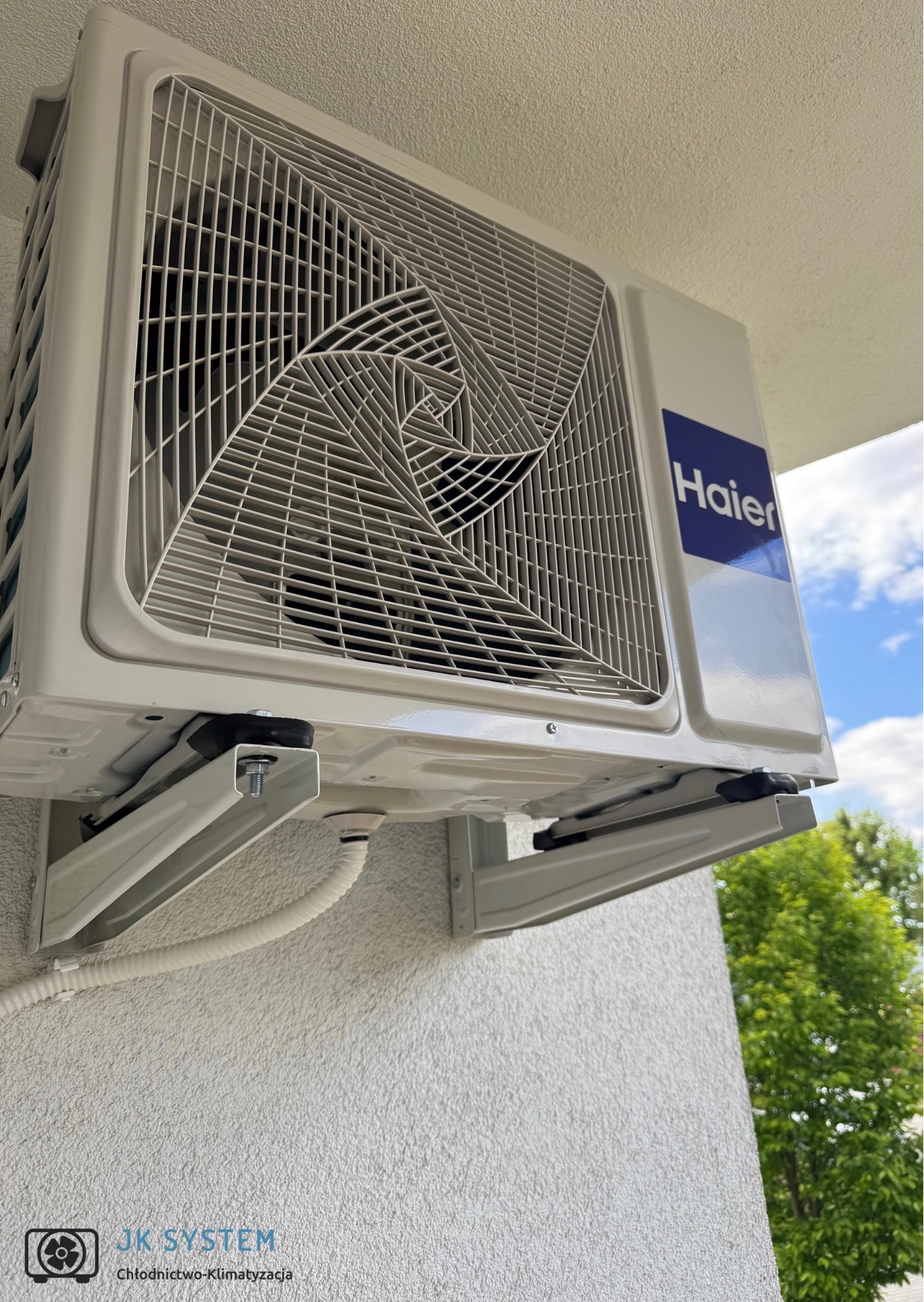 Biały klimatyzator Haier zamontowany na elewacji budynku, widoczny fragment nieba i zieleni. Kątowe ujęcie podkreśla solidność instalacji.