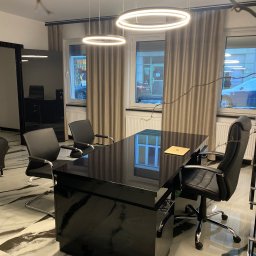 Kancelaria Adwokacka Anna Nowak - Eleganckie biuro: czarne biurko z laptopem, krzesła, marmurowa podłoga i okrągłe lampy sufitowe w jasnym wnętrzu z zasłonami.