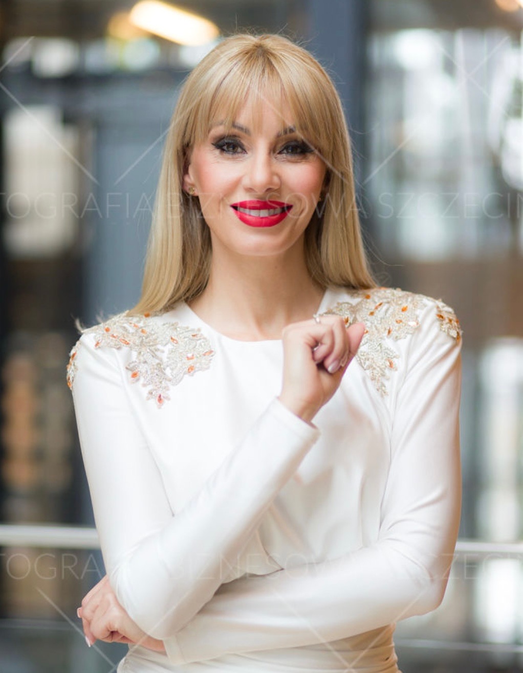 Portret uśmiechniętej kobiety w białej sukience z ozdobnymi ramionami, na rozmytym tle. Czerwona szminka i blond włosy dodają elegancji.