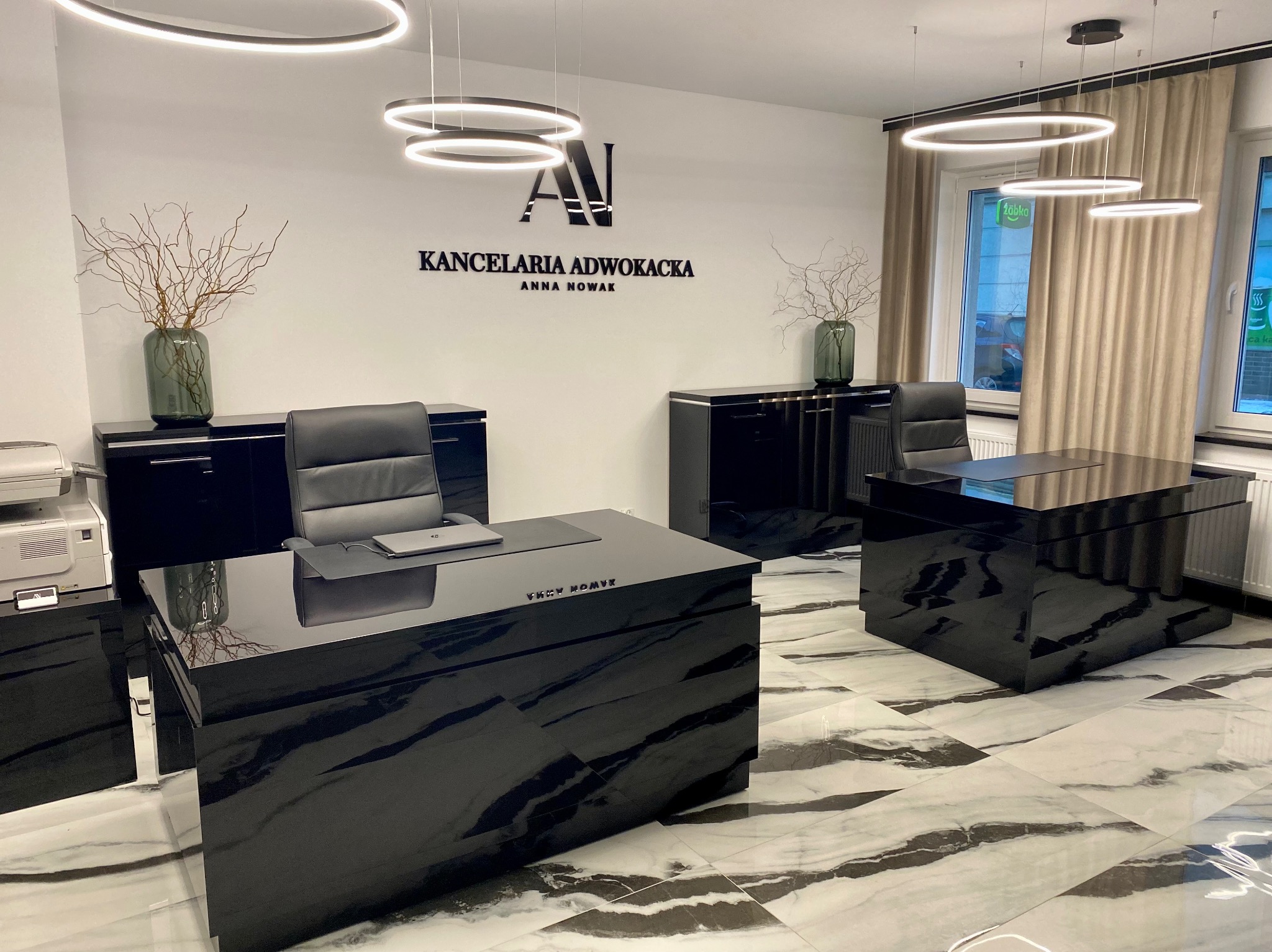 Wnętrze nowoczesnej kancelarii adwokackiej z czarnymi, lakierowanymi meblami, okrągłymi lampami i marmurową podłogą. Na ścianie logo i napis 'Kancelaria Adwokacka Anna Nowak'.