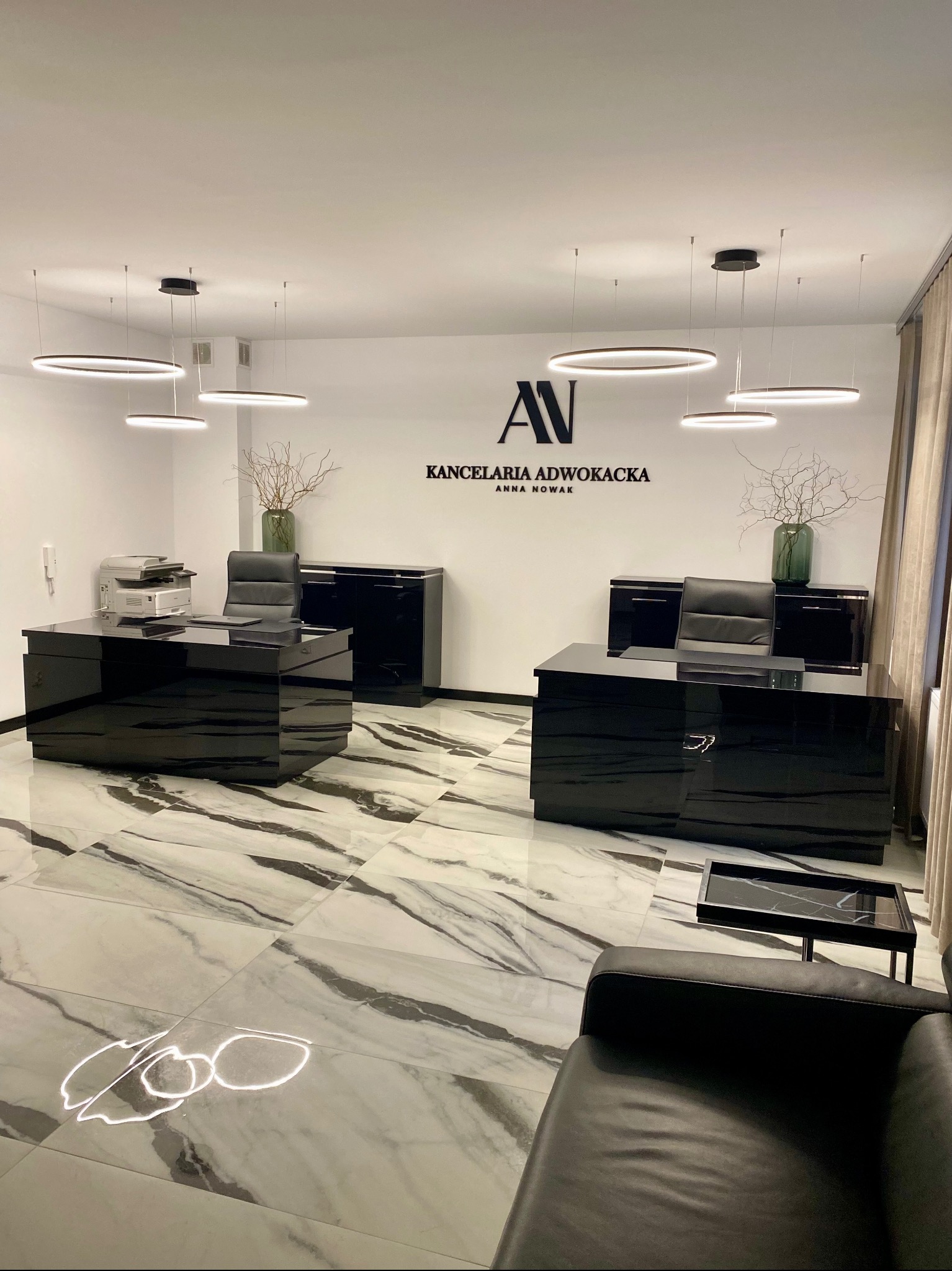 Eleganckie wnętrze kancelarii adwokackiej z czarnymi biurkami, marmurową podłogą i nowoczesnym oświetleniem. Na ścianie logo i nazwa: Kancelaria Adwokacka Anna Nowak.