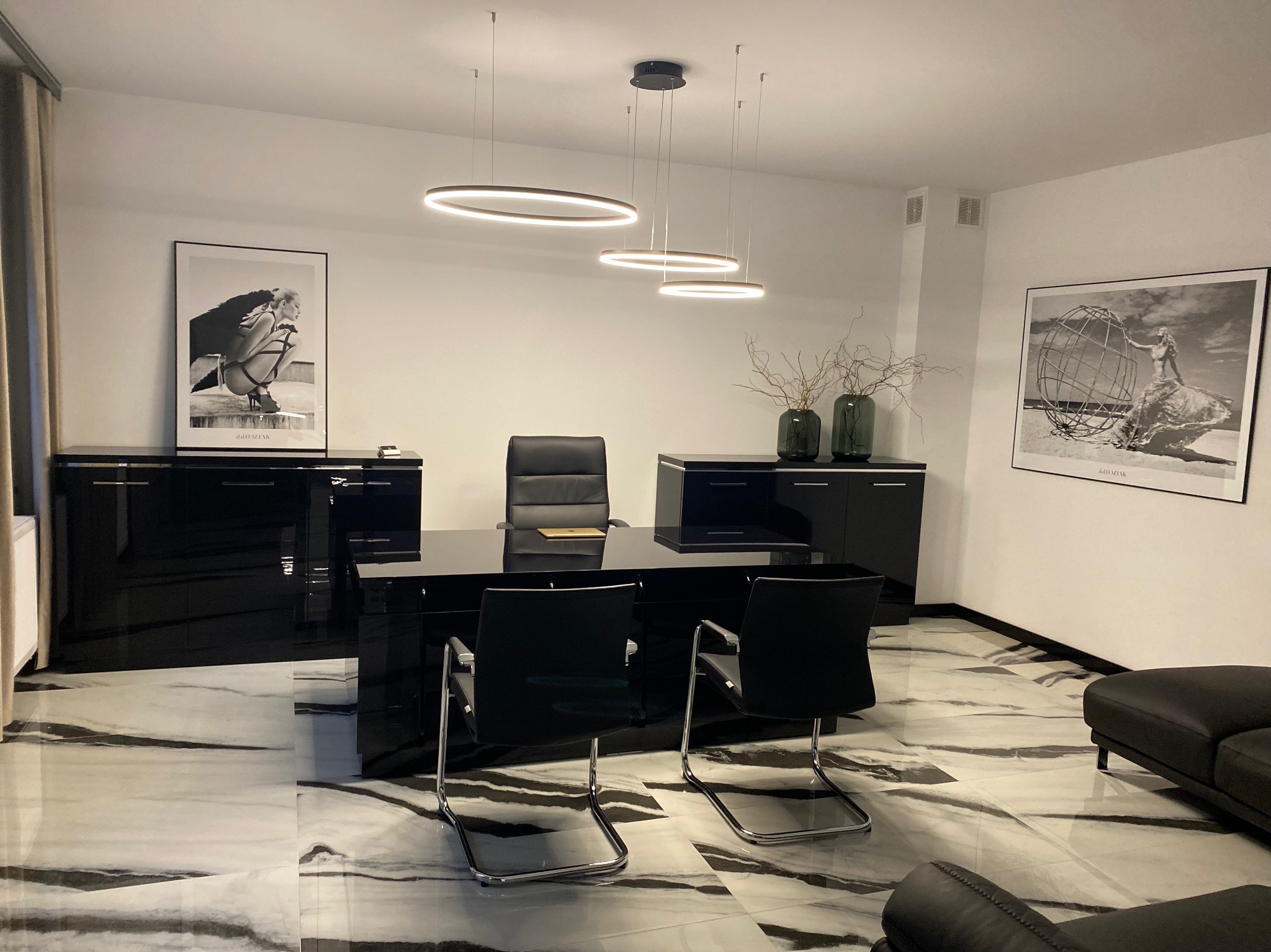 Eleganckie biuro z czarnymi meblami na marmurowej podłodze. Na ścianach wiszą czarno-białe grafiki. Nad biurkiem nowoczesna lampa pierścieniowa.