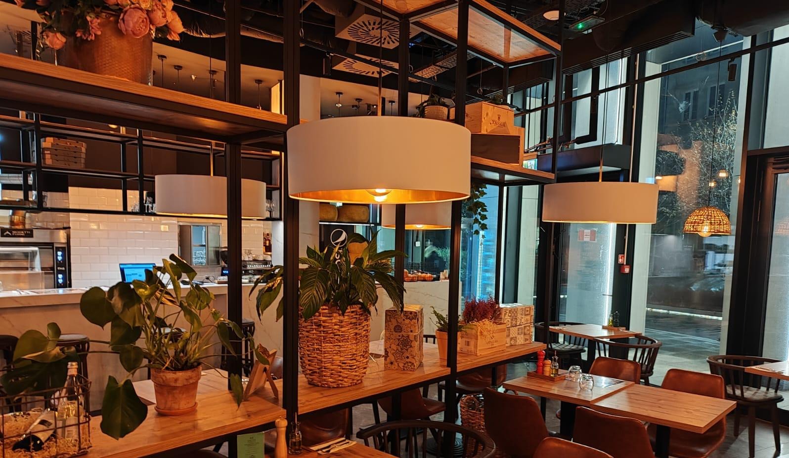 Nowoczesne wykończenie wnętrza restauracji: drewniane półki z roślinami, lampy z białymi abażurami i widok na kuchnię z białymi płytkami.