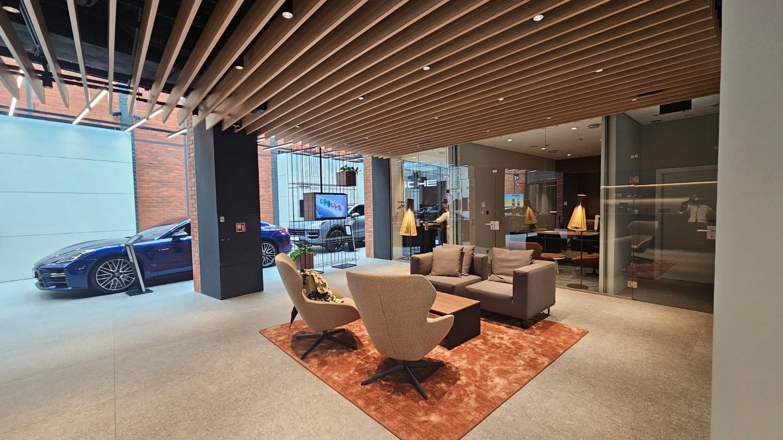 Nowoczesne wnętrze biura z drewnianym sufitem, ceglaną ścianą i luksusowymi samochodami. Widoczne meble wypoczynkowe i przeszklenia, tworzące elegancką przestrzeń.