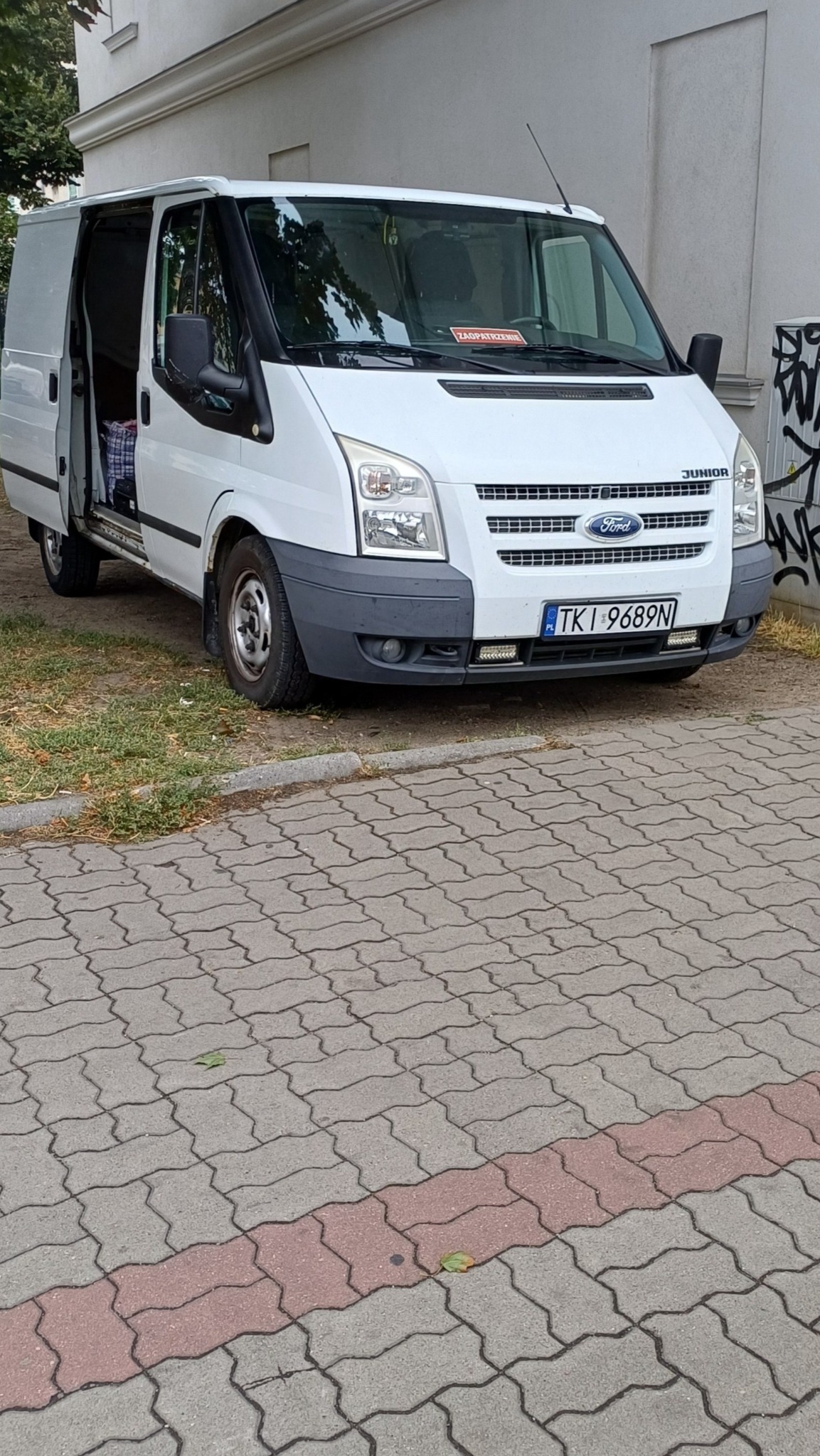 Biały Ford Transit Junior z otwartymi drzwiami bocznymi, zaparkowany na chodniku z kostki. Widoczna tablica rejestracyjna i naklejka 'Zaopatrzenie' na przedniej szybie.