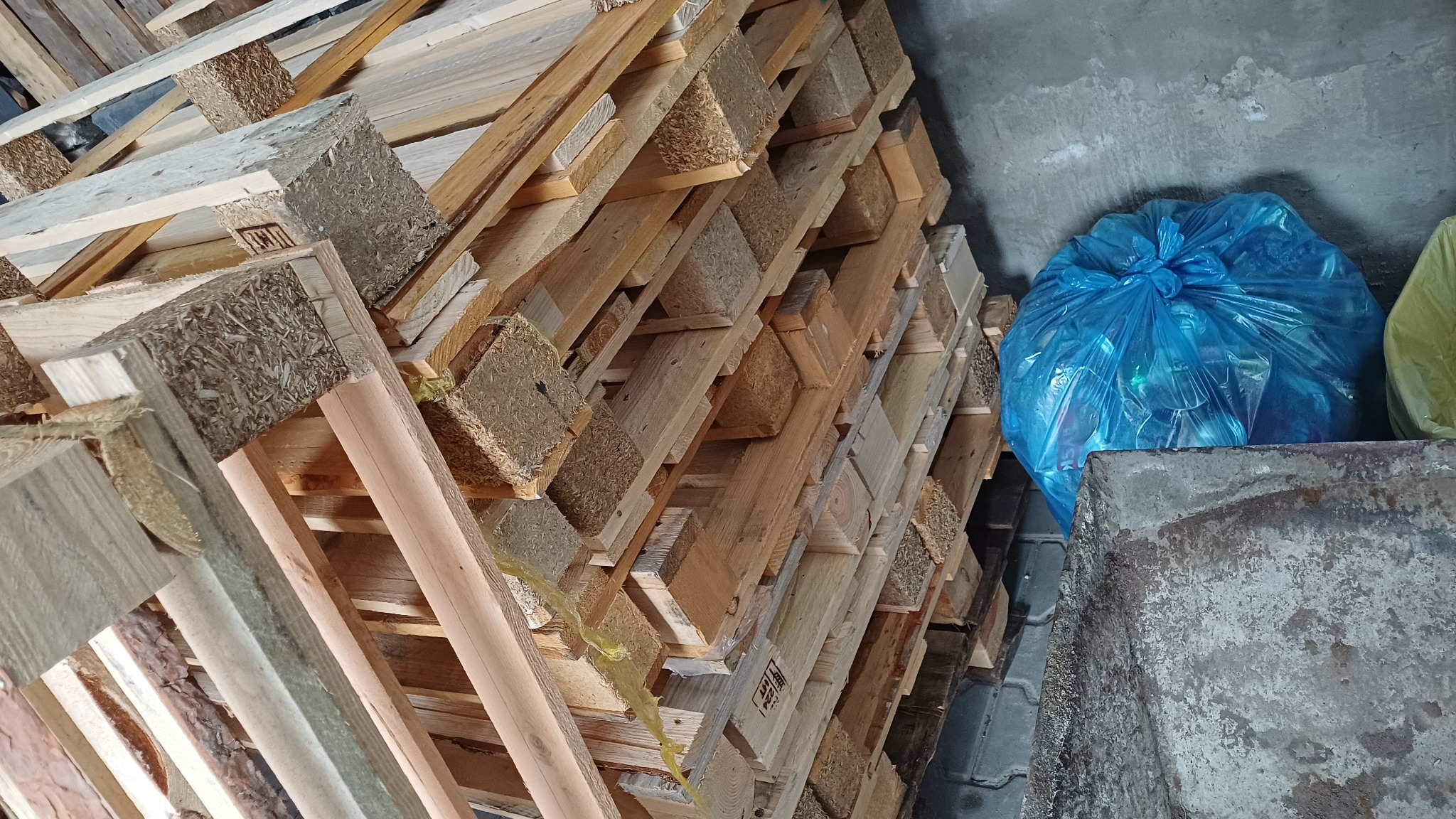 Stos drewnianych palet ułożonych pod kątem, obok niebieski worek na śmieci i metalowa skrzynia. Magazynowe otoczenie, szara ściana w tle.