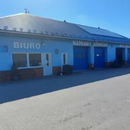 Niebieski budynek warsztatu samochodowego z napisem 'Biuro', 'Naprawy serwisowe' i 'Wulkanizacja'. Przed budynkiem zaparkowany samochód dostawczy i opony.