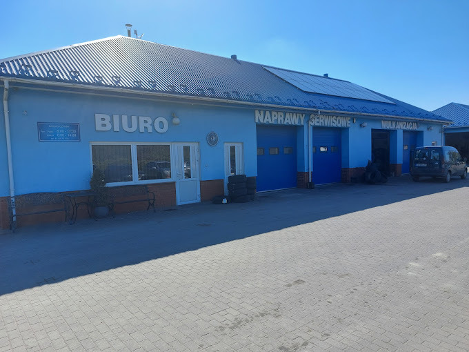 Niebieski budynek warsztatu samochodowego z napisem 'Biuro', 'Naprawy serwisowe' i 'Wulkanizacja'. Przed budynkiem zaparkowany samochód dostawczy i opony.
