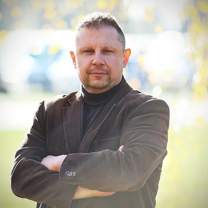 Portret pewnego mężczyzny w brązowej marynarce sztruksowej, z założonymi rękami, na rozmytym tle jesiennych drzew.