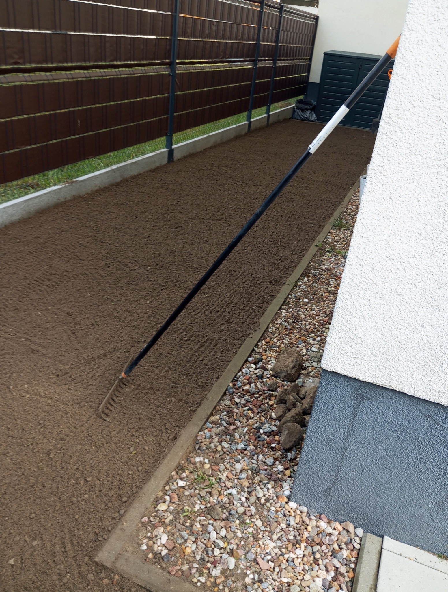 Przygotowany grunt pod trawnik z widocznymi śladami grabi, obrzeżem z kostki i ogrodzeniem panelowym w tle. Widoczne kamienie i fragment elewacji budynku.