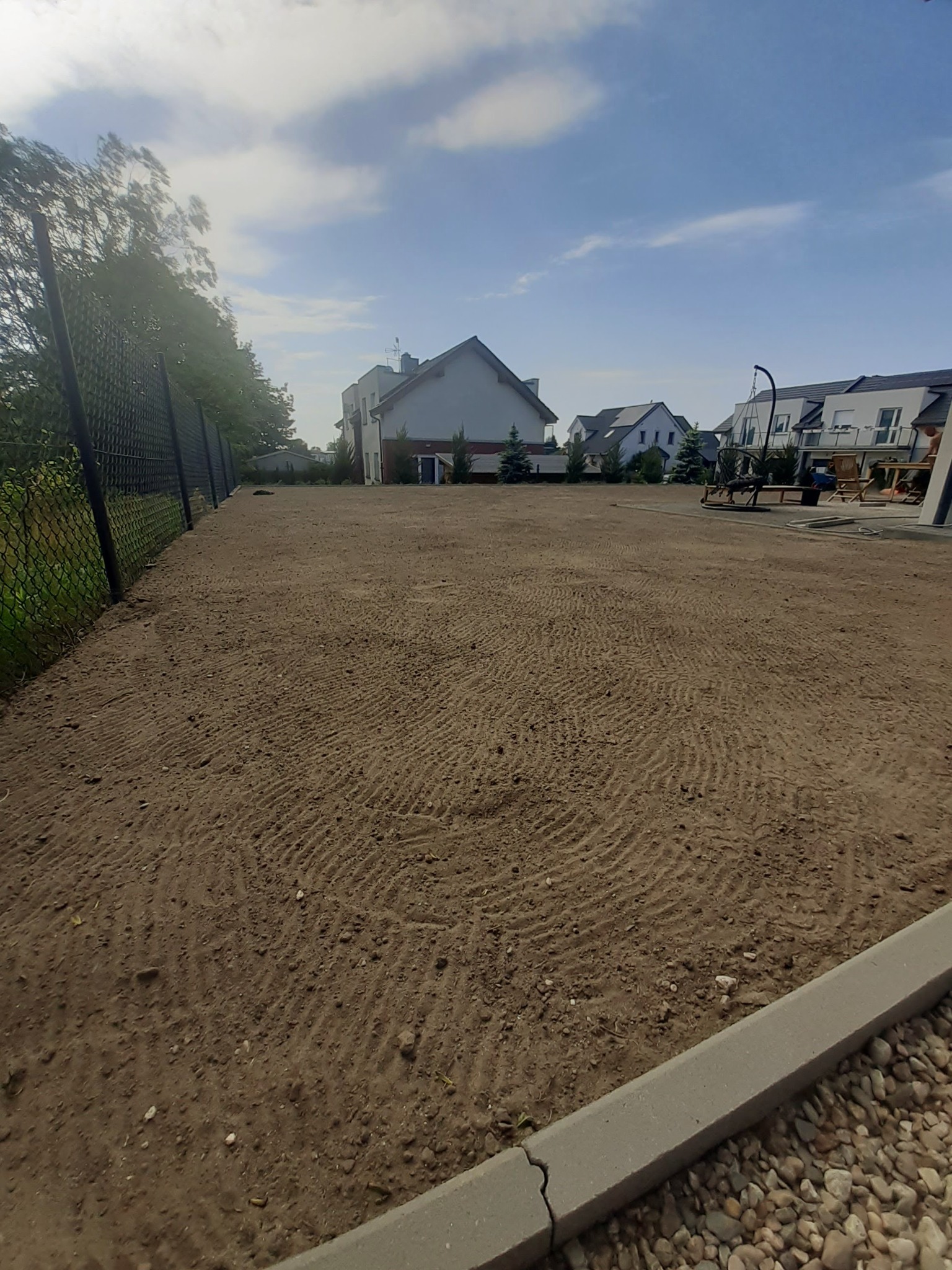 Przygotowany teren pod trawnik z widocznymi śladami po bronowaniu, ogrodzony siatką. W tle zabudowa osiedlowa i błękitne niebo.