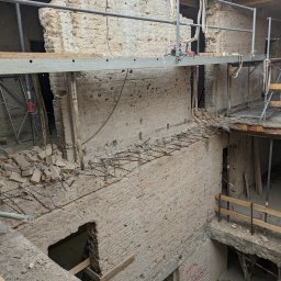 Pro-mon - Częściowo wyburzona ceglana ściana z widocznym zbrojeniem i narzędziami na rusztowaniu. W tle żółty kask budowlany. Prace wyburzeniowe w toku.