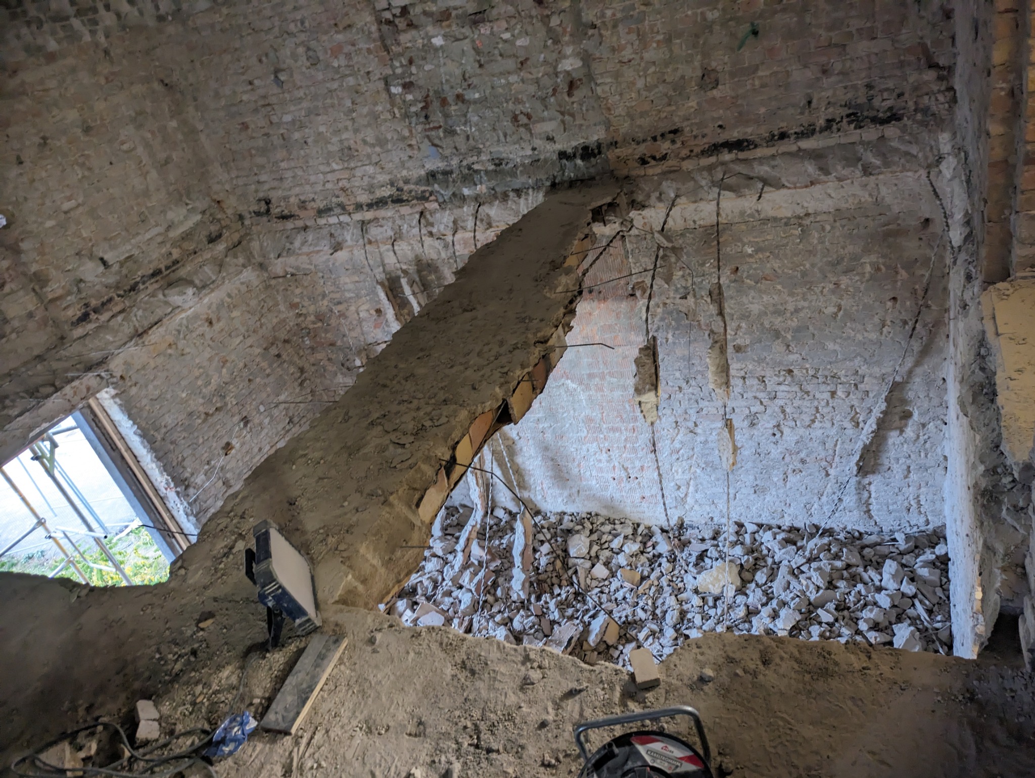 Częściowo wyburzona ceglana ściana z widocznym zbrojeniem i gruzem, ukazująca postęp prac rozbiórkowych wewnątrz budynku. Widoczne narzędzia i oświetlenie.