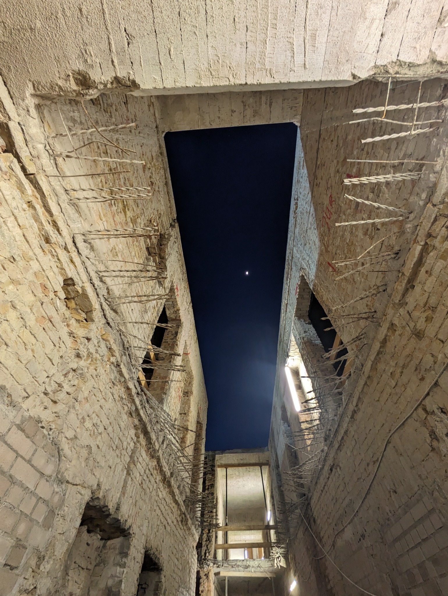 Wnętrze budynku w trakcie rozbiórki, widok na nocne niebo z księżycem przez otwór w stropie. Fragmenty cegieł i wystające zbrojenia. Ujęcie z dołu.