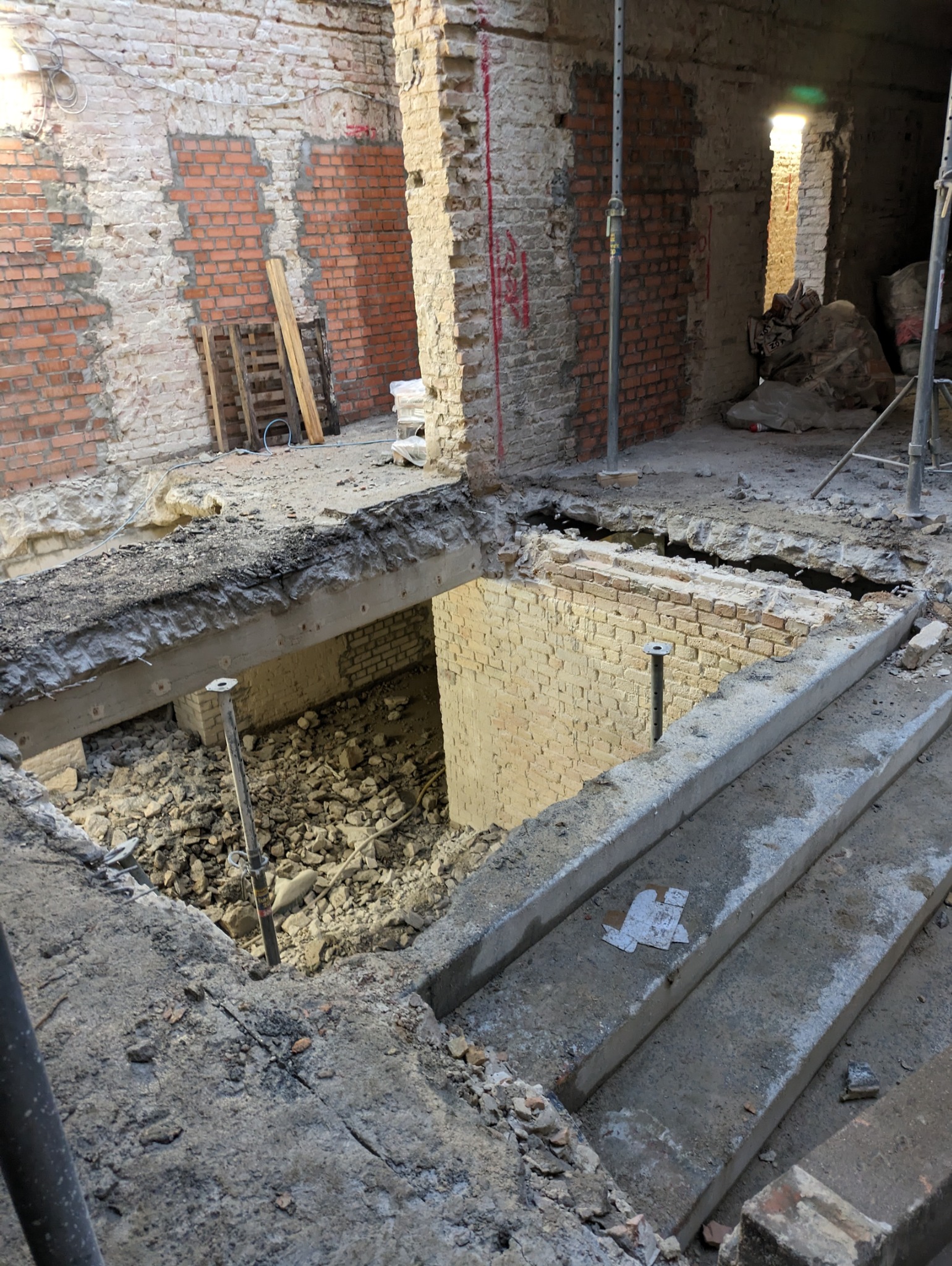 Wnętrze w trakcie rozbiórki: odsłonięte cegły, fragmenty betonu i gruzu. Widoczne podpory i elementy konstrukcyjne. Otwór w podłodze ukazuje wnętrze piwnicy.