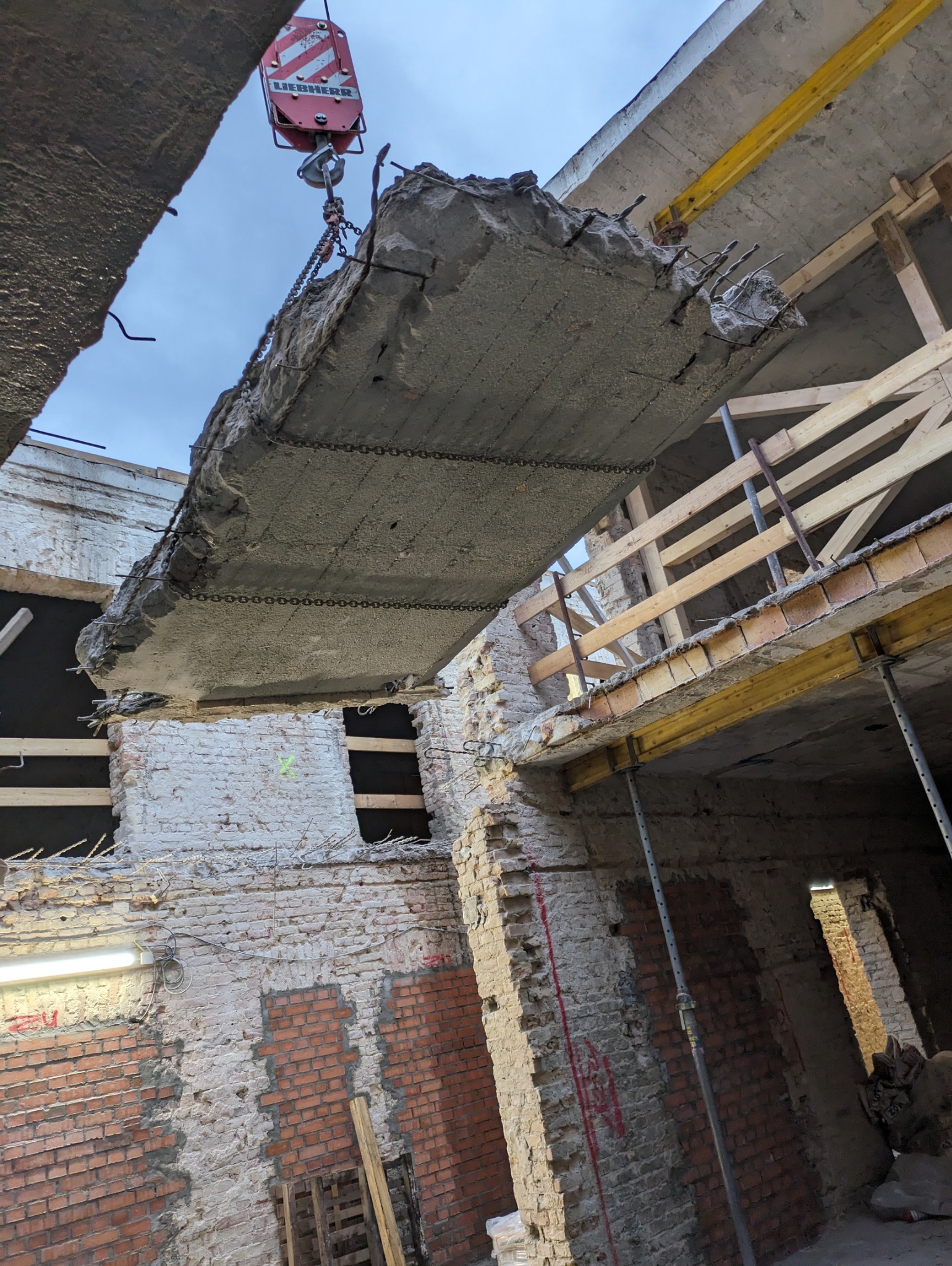 Demontaż stropu gruzobetonowego za pomocą dźwigu w budynku z odsłoniętą cegłą. Widoczne zbrojenia i podpory stropu.