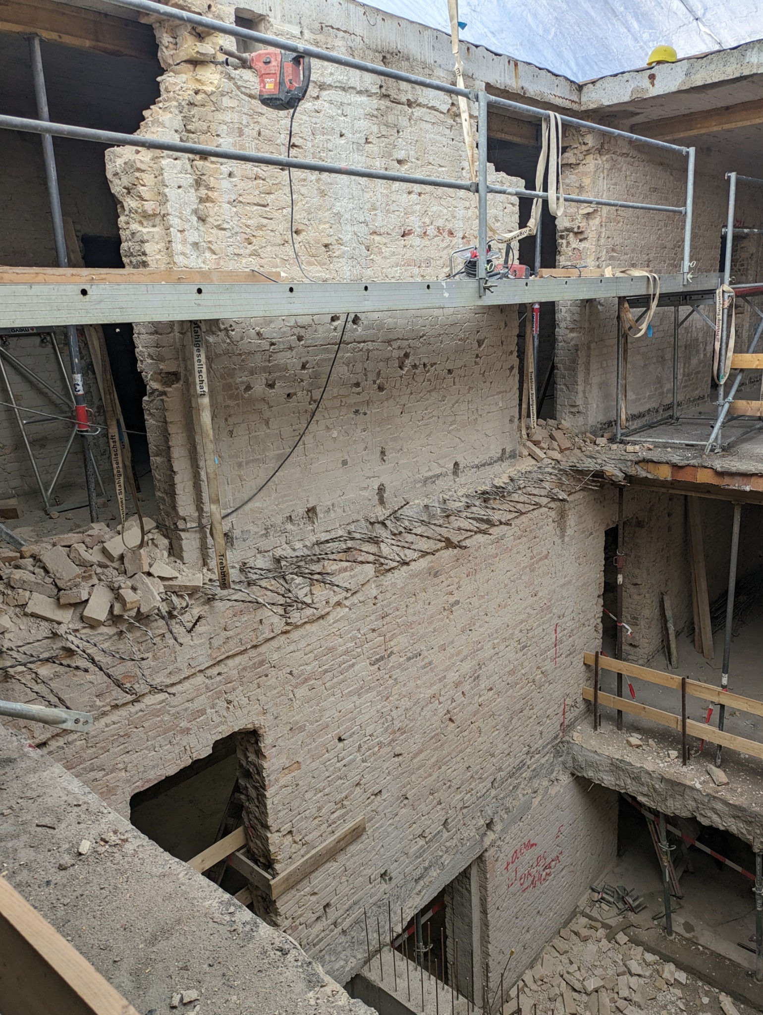 Częściowo wyburzona ceglana ściana z widocznym zbrojeniem i narzędziami na rusztowaniu. W tle żółty kask budowlany. Prace wyburzeniowe w toku.