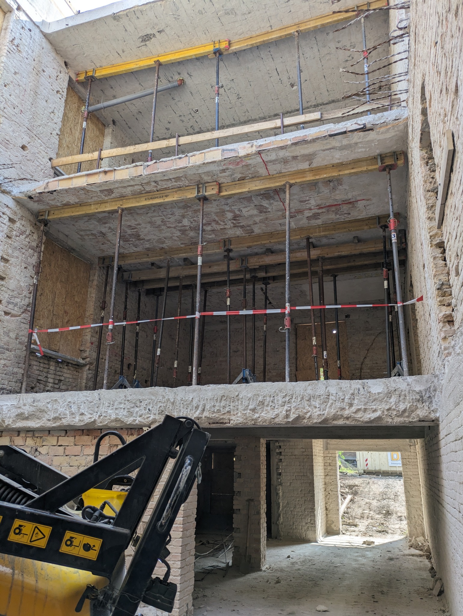 Wnętrze budynku w trakcie rozbiórki, widoczne metalowe podpory, fragment maszyny budowlanej i gruz. Ceglane ściany z widocznymi uszkodzeniami.