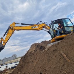 Zdun-Bud - Żółta koparka JCB na wzgórzu ziemi z operatorem w kabinie, w tle budynek i niebo z chmurami. Wykop pod fundamenty w tle.