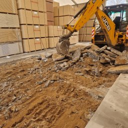 Zdun-Bud - Żółta koparka JCB w hali z ziemią i gruzem, na tle palet z płytami Swiss Krono. Praca w toku, dobrze oświetlone wnętrze, widoczny numer kontaktowy na maszynie.
