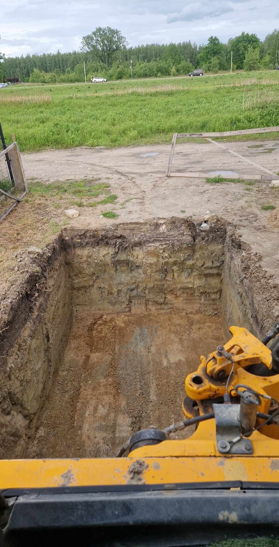 Wykopany dół w ziemi z widoczną częścią żółtej maszyny budowlanej, prawdopodobnie koparki. W tle łąka i drzewa pod pochmurnym niebem.