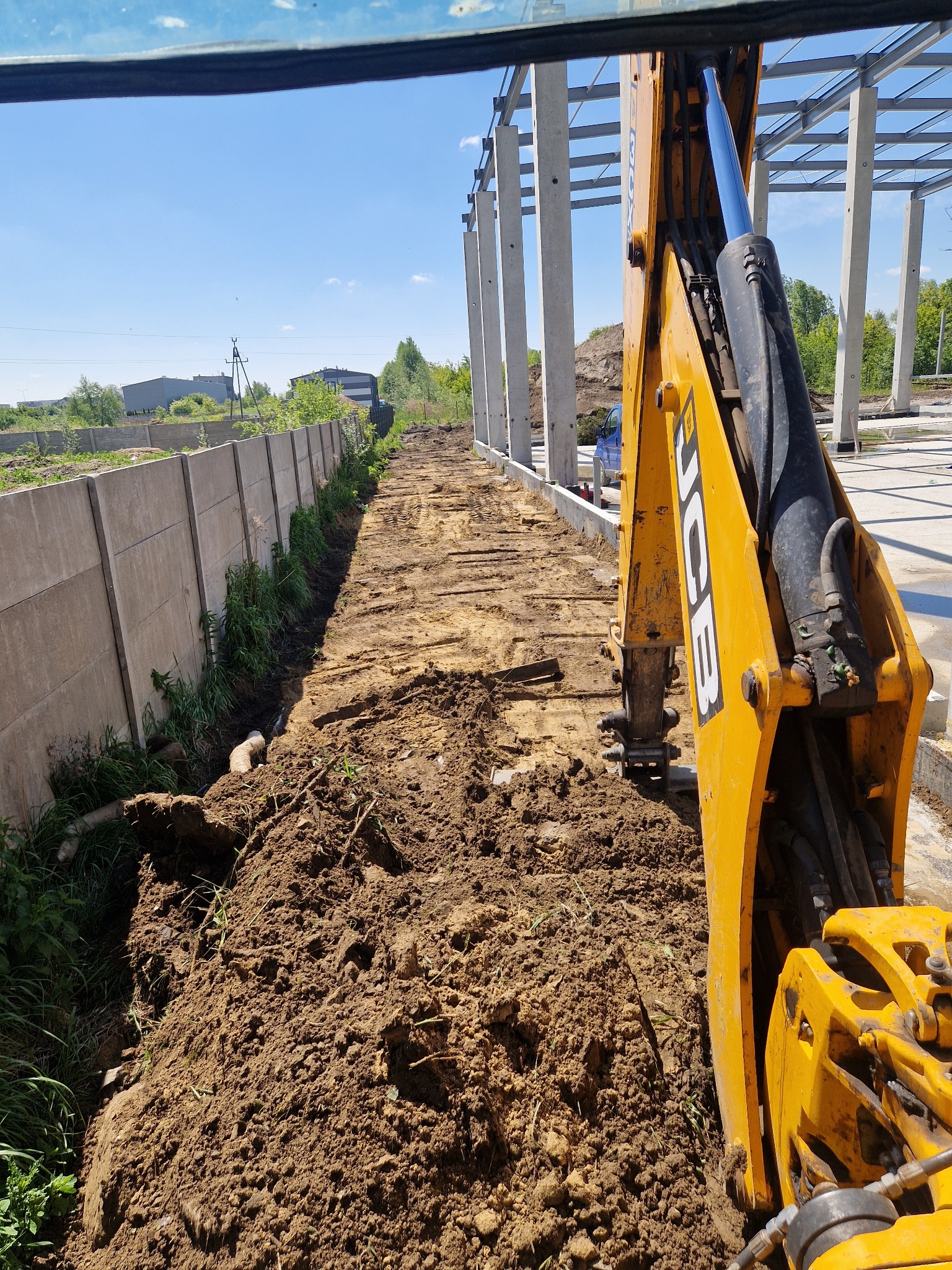Żółta koparka JCB w trakcie prac ziemnych na placu budowy, widok z kabiny. W tle betonowe ogrodzenie i konstrukcja hali.