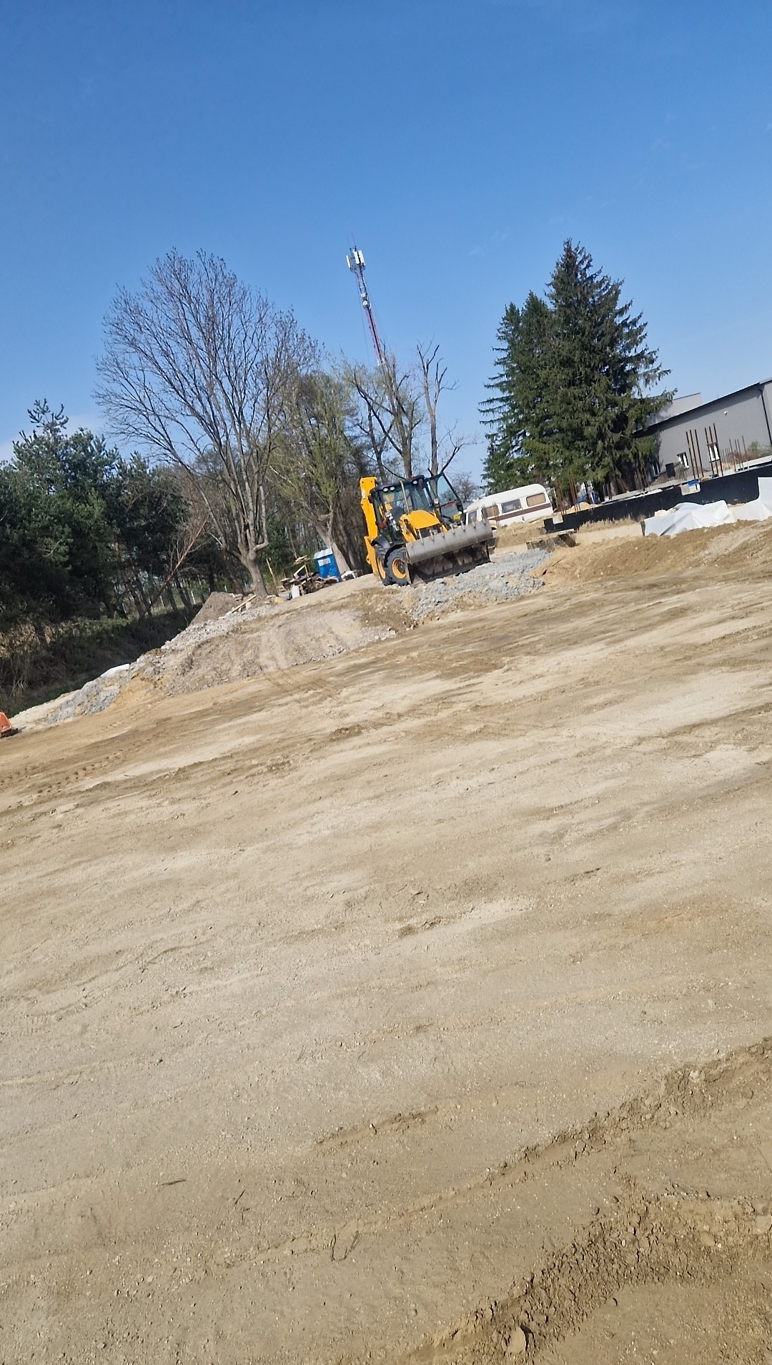 Żółta koparko-ładowarka na placu budowy w słoneczny dzień. Widoczne drzewa, przyczepa kempingowa i maszt telekomunikacyjny w tle. Ślady opon na piasku.