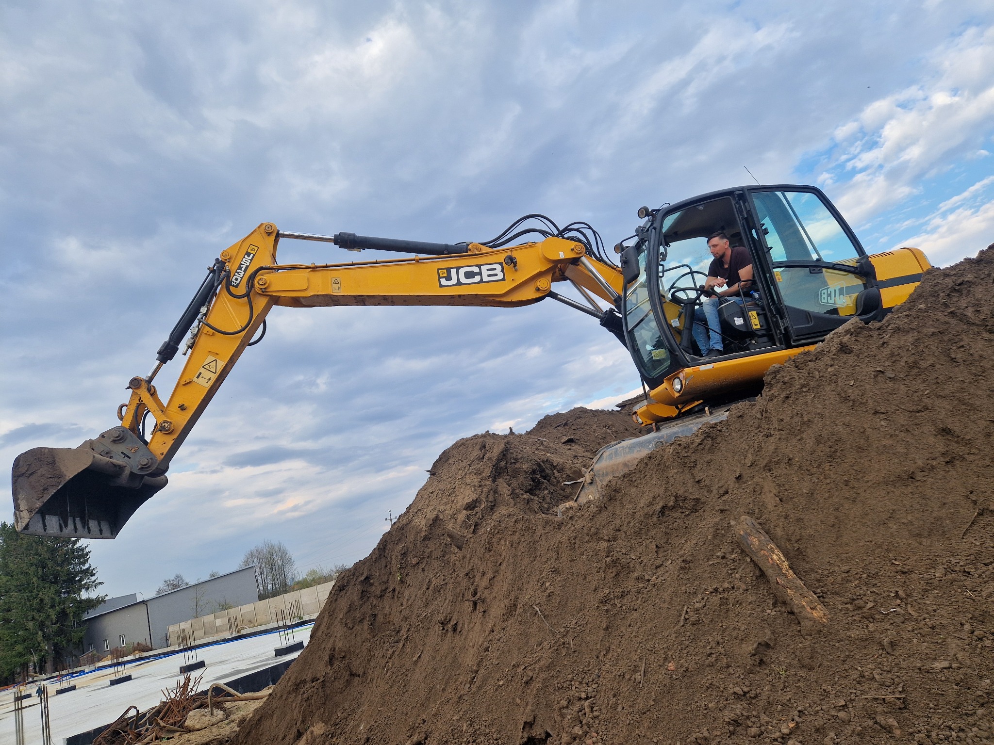 Żółta koparka JCB na wzgórzu ziemi z operatorem w kabinie, w tle budynek i niebo z chmurami. Wykop pod fundamenty w tle.