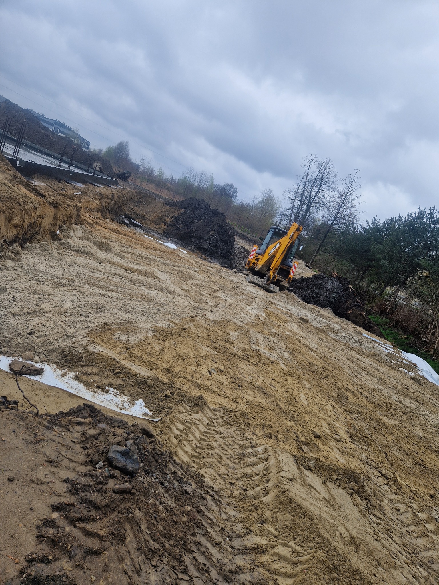 Żółta koparka JCB na placu budowy, przygotowany teren pod fundamenty. Widoczne ślady opon na piasku i czarna ziemia w tle. Pochmurne niebo.