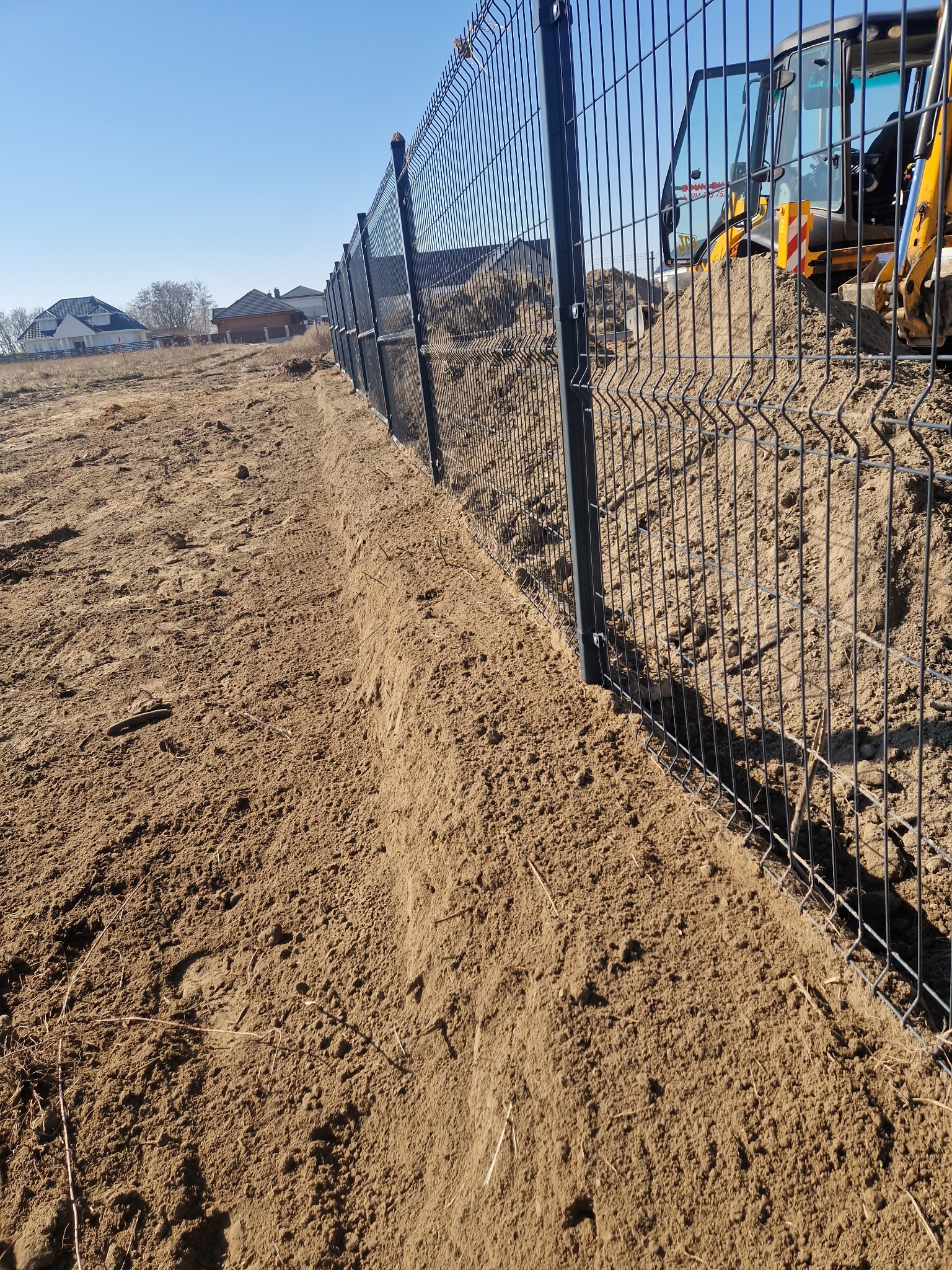Montaż ogrodzenia panelowego na przygotowanym terenie. W tle widoczna koparka, sugerująca prace ziemne. Słoneczny dzień, błękitne niebo. W tle zabudowa jednorodzinna.