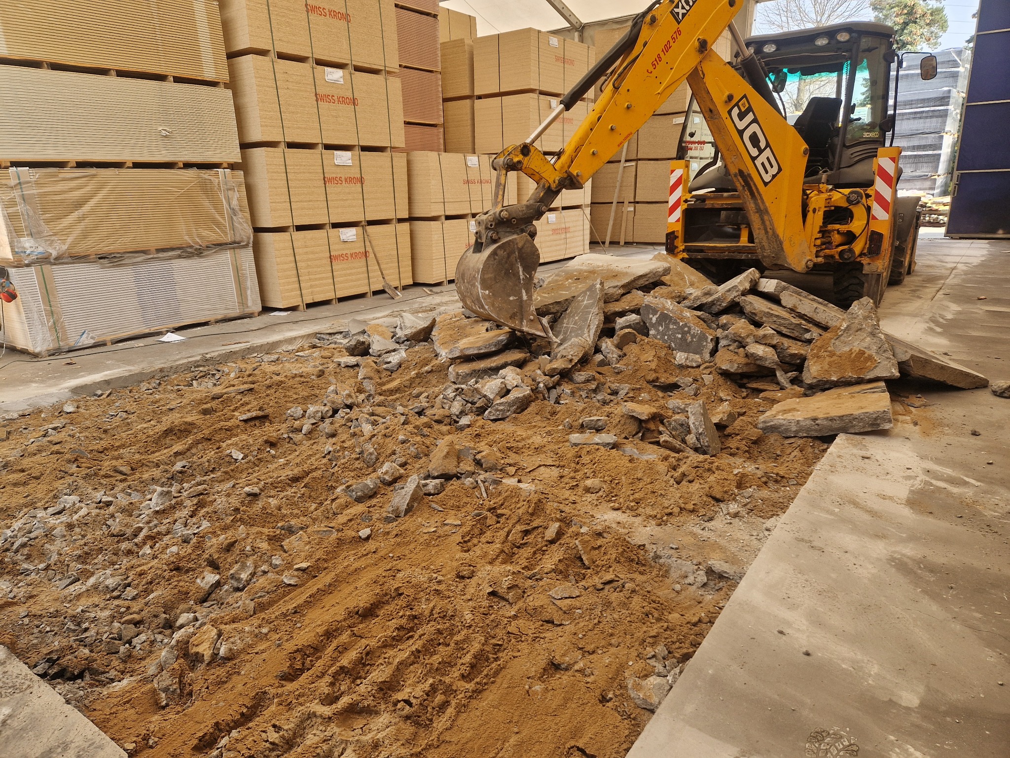 Żółta koparka JCB w hali z ziemią i gruzem, na tle palet z płytami Swiss Krono. Praca w toku, dobrze oświetlone wnętrze, widoczny numer kontaktowy na maszynie.