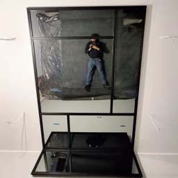 CROMO JMI Rozalia Milbrod - Wnętrze z dużym lustrem w czarnej ramie, w którym odbija się osoba robiąca zdjęcie telefonem. Widoczne kable wystające ze ściany i elementy przygotowania do montażu.