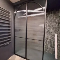 Nowoczesne drzwi szklane z czarną ramą i półokrągłym wykończeniem, widoczne na tle wzorzystej ściany i grzejnika żeberkowego. Nietypowe wykończenie szkła.