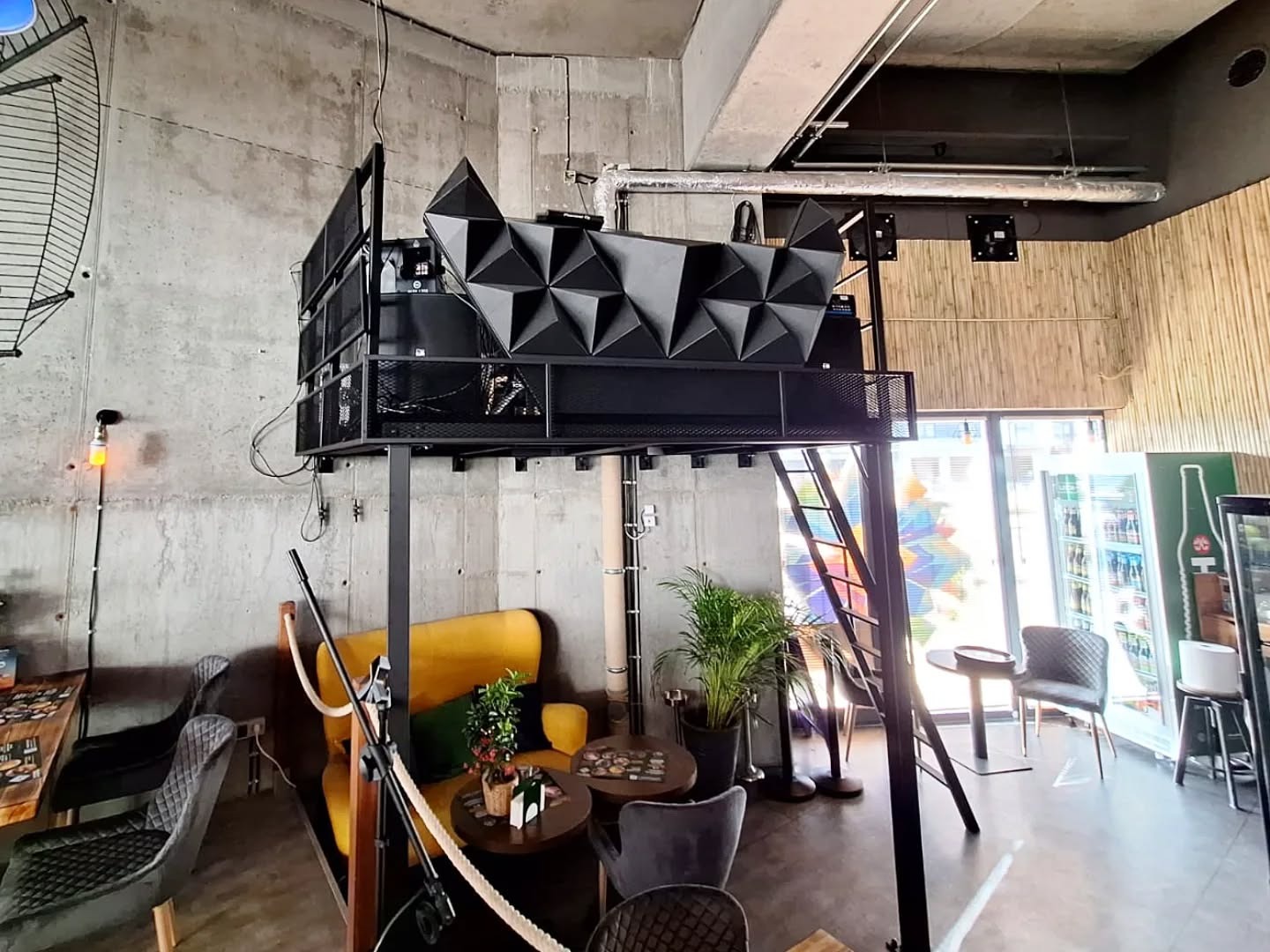 Metalowa konstrukcja z platformą w loftowym wnętrzu kawiarni. Schody prowadzą na górę, gdzie znajduje się czarny, geometryczny element dekoracyjny.