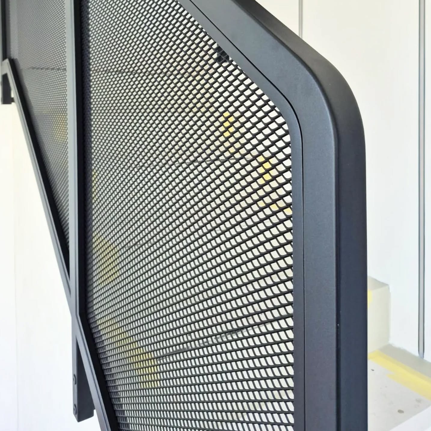 Czarna, metalowa balustrada schodowa z siatką o drobnych oczkach, zamontowana na białej ścianie. Widoczny fragment schodów z żółtą taśmą.