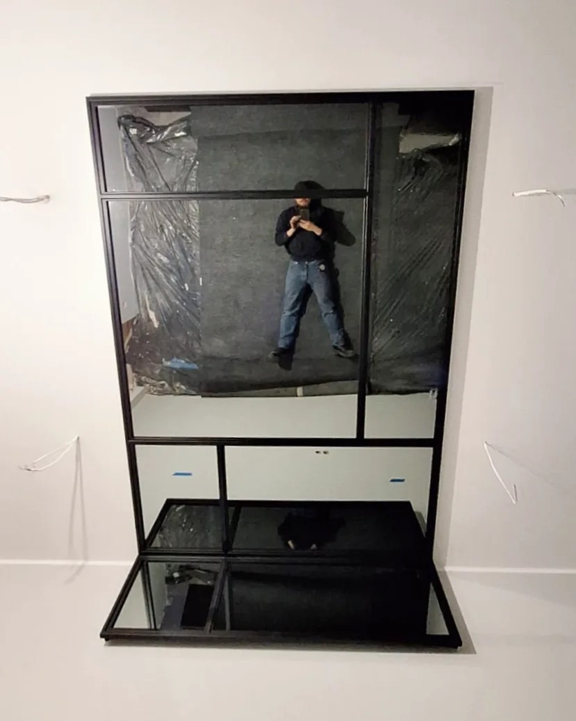 Wnętrze z dużym lustrem w czarnej ramie, w którym odbija się osoba robiąca zdjęcie telefonem. Widoczne kable wystające ze ściany i elementy przygotowania do montażu.