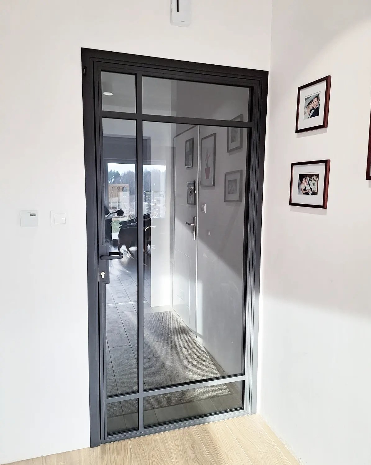 Szklane drzwi w czarnej ramie z podziałami, widok z korytarza na wnętrze domu. Na ścianie wiszą ramki ze zdjęciami.
