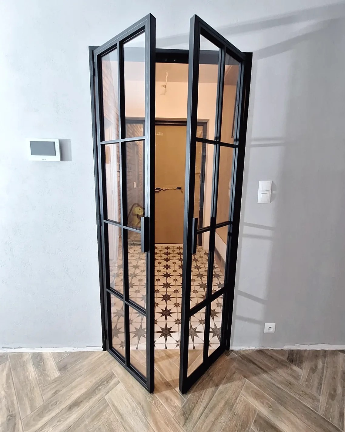 Otwarta, dwuskrzydłowa metalowa ścianka działowa z szybami, pomalowana na czarno, oddzielająca dwa pomieszczenia wykończone w różnym stylu.