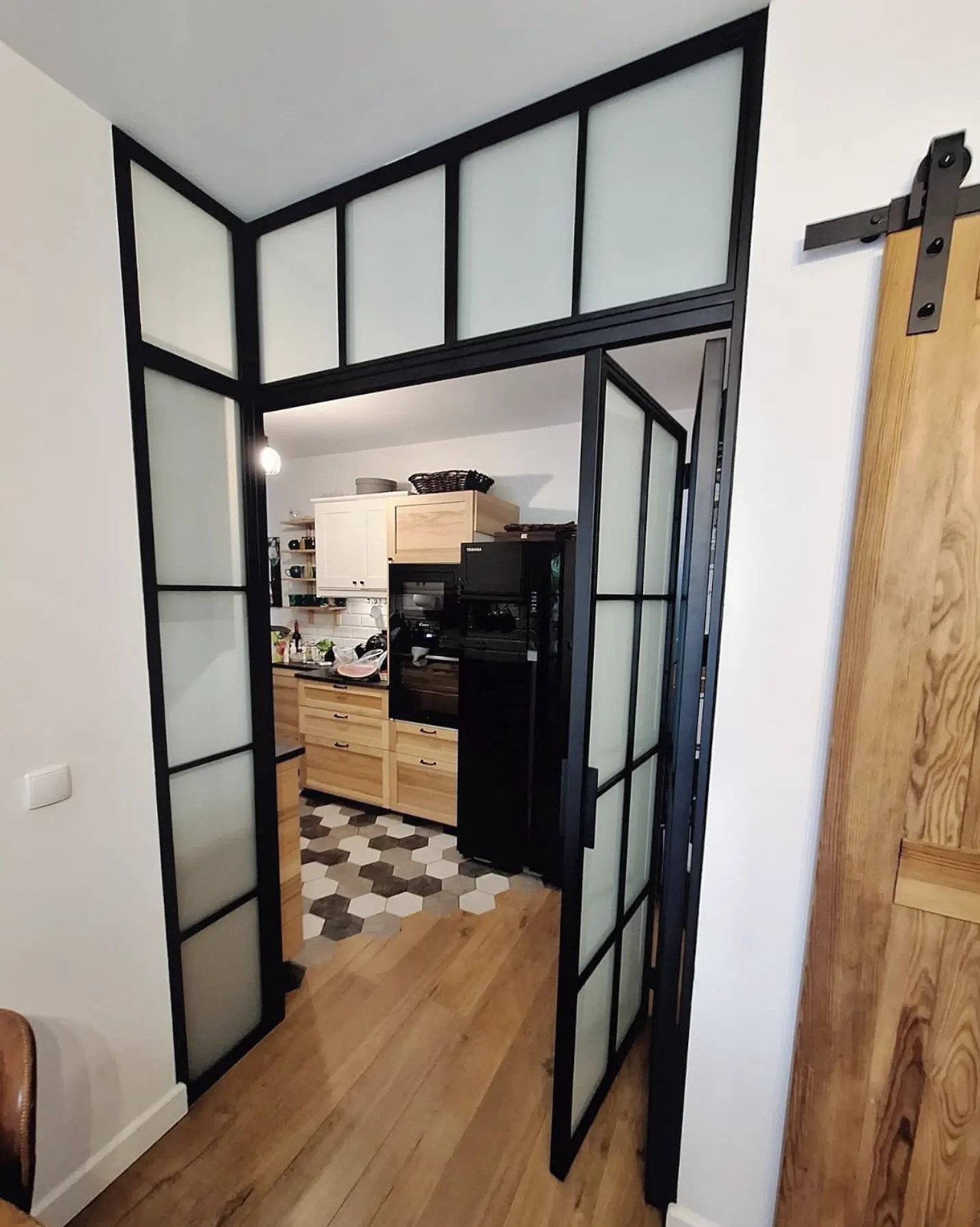 Nowoczesne drzwi szklane z czarnymi szprosami oddzielają kuchnię od salonu. Widoczny fragment drewnianych drzwi przesuwnych i heksagonalna podłoga w kuchni.