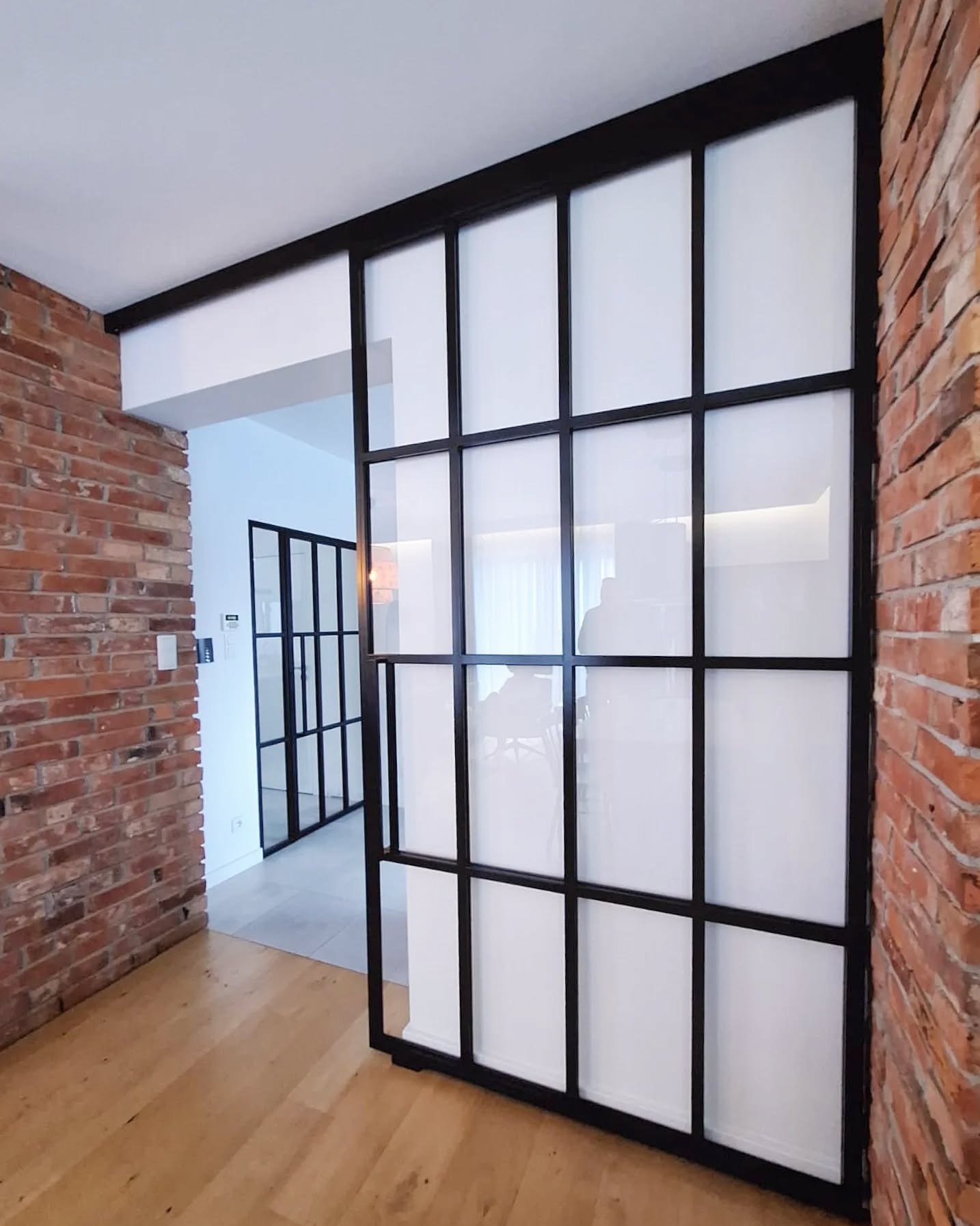Loftowe drzwi przesuwne z czarną ramą i mlecznym szkłem, ustawione na tle ściany z czerwonej cegły. Widoczny fragment podłogi z jasnego drewna.