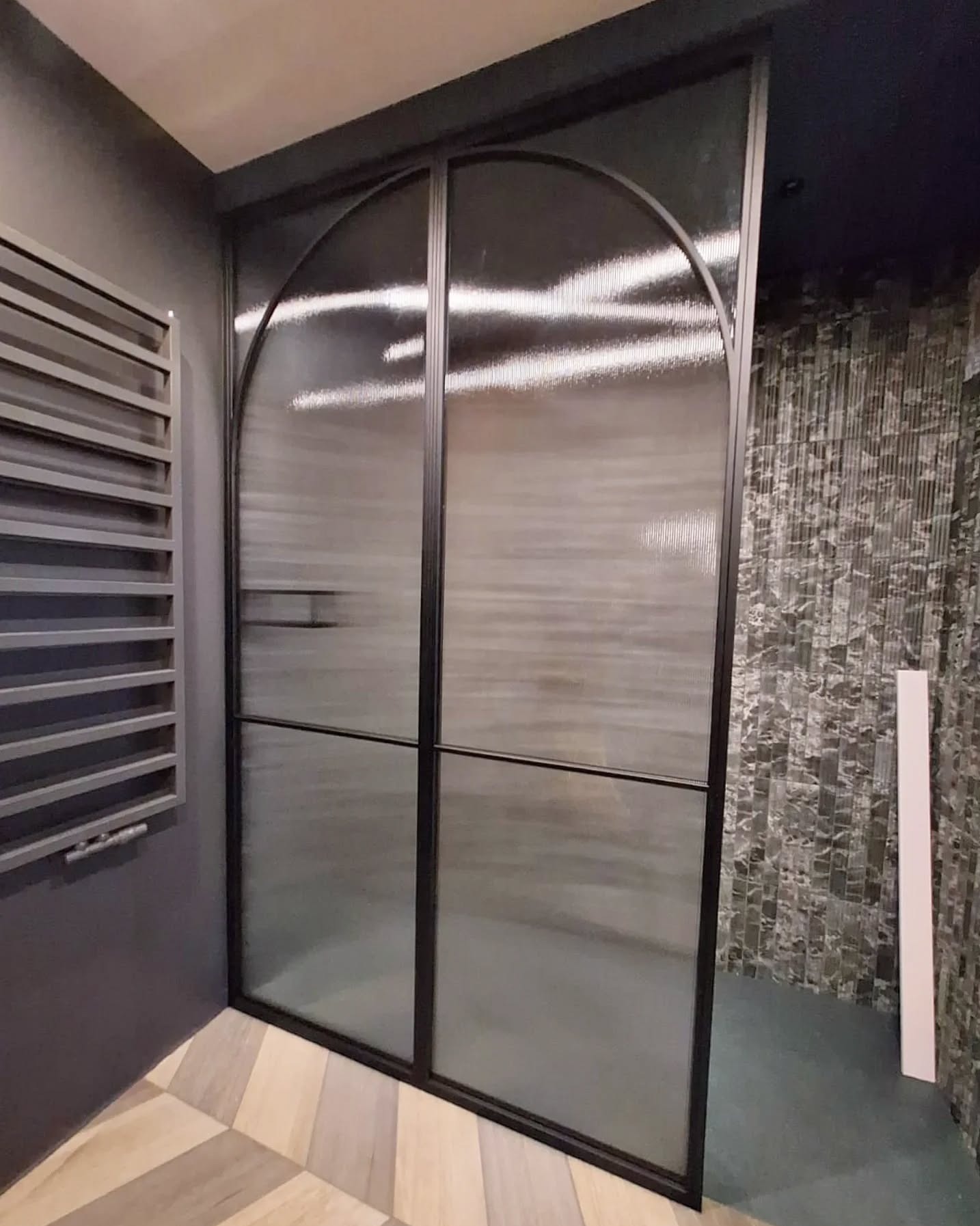 Nowoczesne drzwi szklane z czarną ramą i półokrągłym wykończeniem, widoczne na tle wzorzystej ściany i grzejnika żeberkowego. Nietypowe wykończenie szkła.